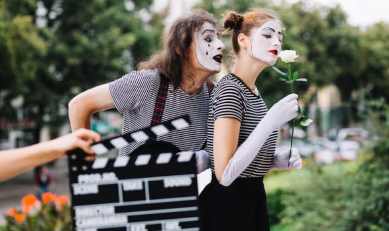 Dve odrasle glumice sa belom scenskom šminkom izvode pantomimu na otvorenom, dok se u prvom planu nalazi filmska klapa